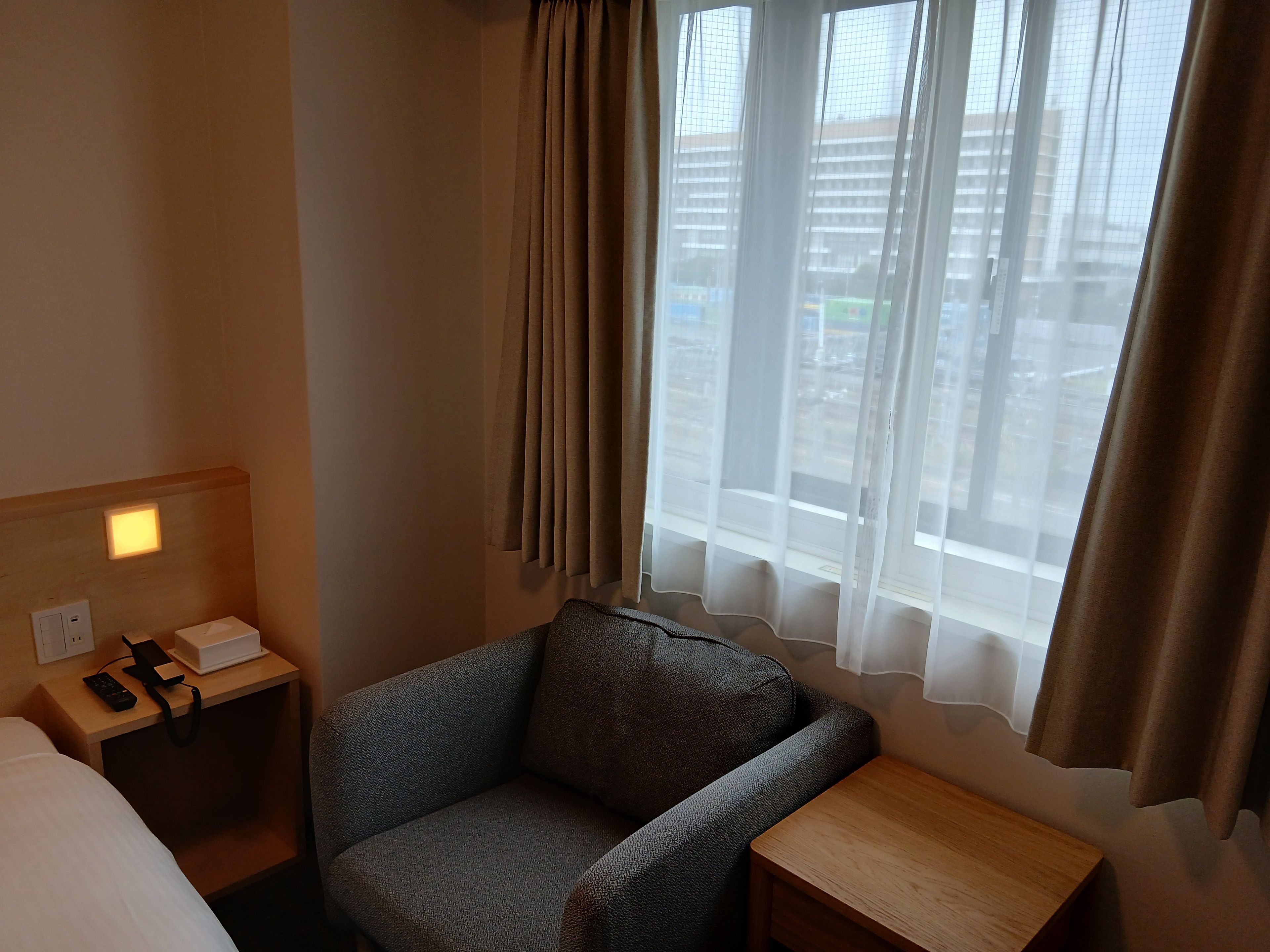 A hotel room with a bed, a small table, a chair, a nightstand with a phone and remote, and a window with curtains.