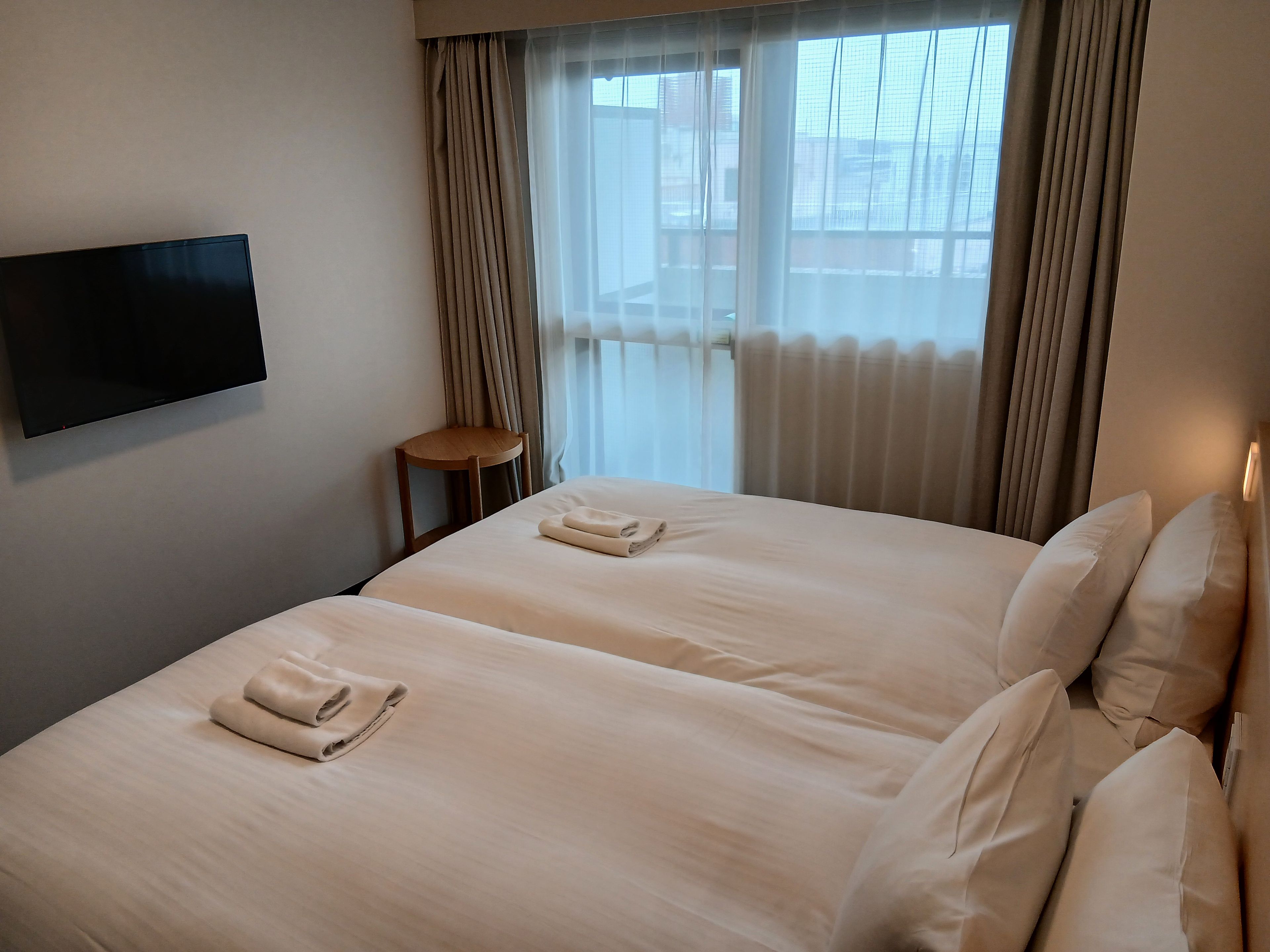 Habitació d'hotel amb llit, una cadira, televisió i finestra amb cortines.