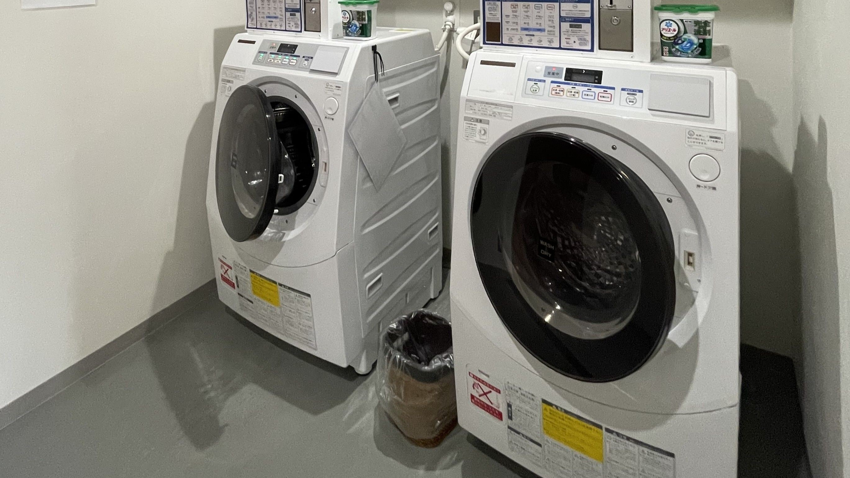 Two front-loading washing machines with control panels, a trash bin, and a wall-mounted control panel with instructions.
