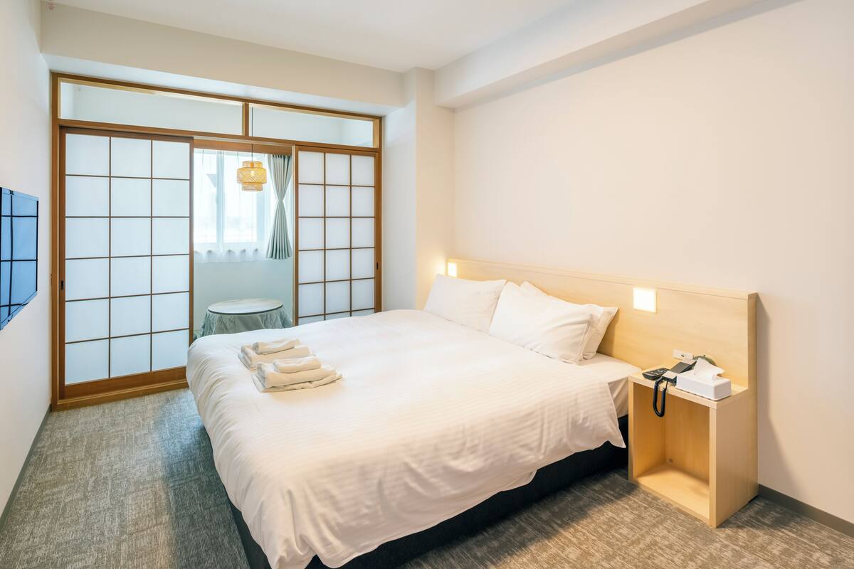 Quarto de hotel com uma cama grande, uma mesa de cabeceira com telefone e um objeto, uma pequena mesa redonda e uma porta de tela deslizante estilo shoji.