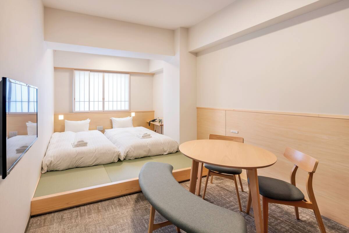 Quarto de hotel com duas camas, uma mesinha e um banquinho.