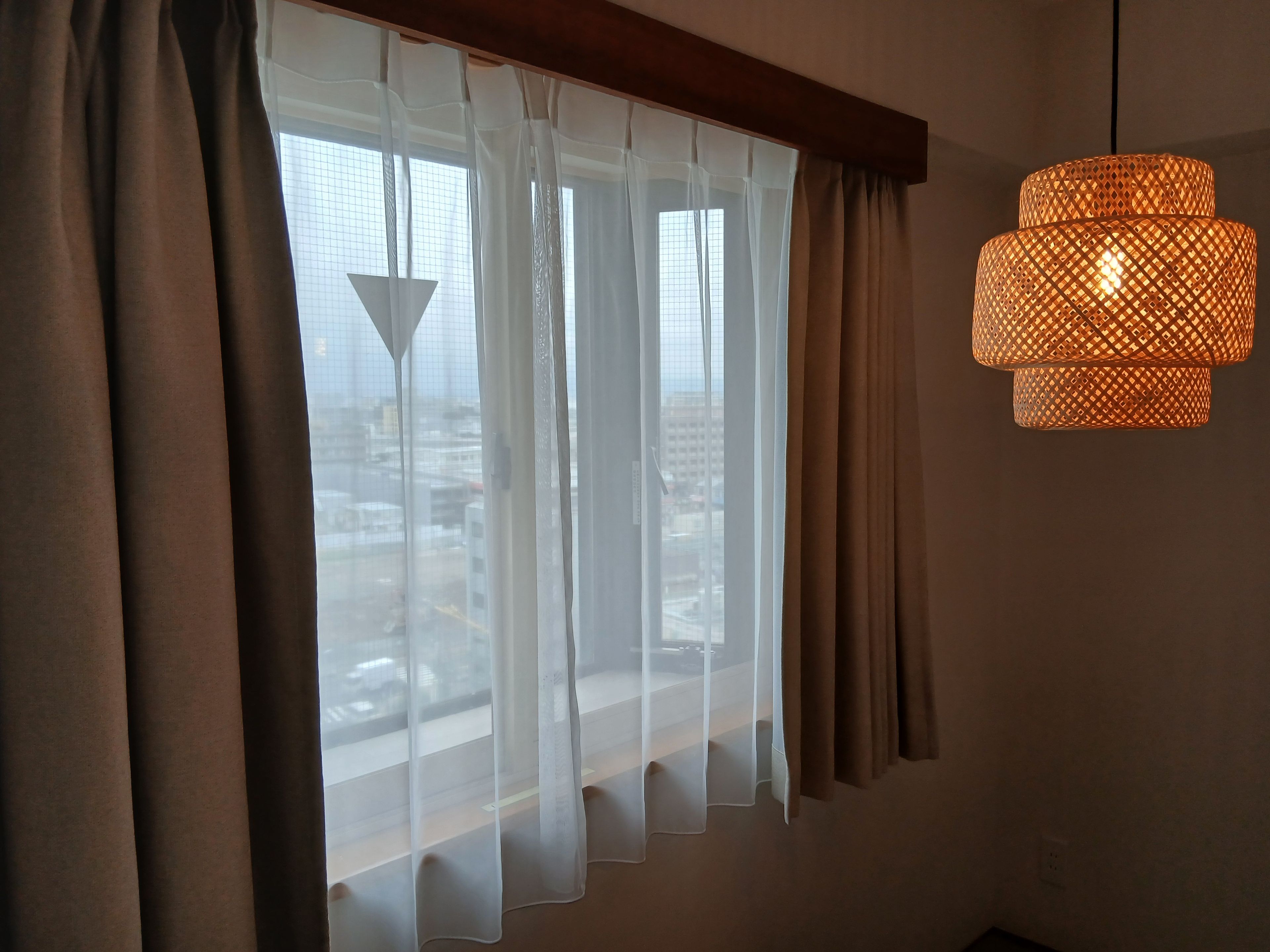 Ett rum med ett stort fönster, tunna, genomskinliga gardiner och en hängande lampa med flätad skärm.