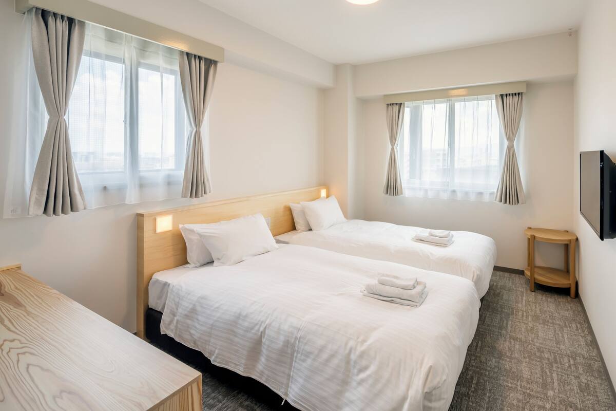 Quarto de hotel com duas camas, uma escrivaninha de madeira, uma pequena mesa de cabeceira e uma televisão de tela plana presa à parede.