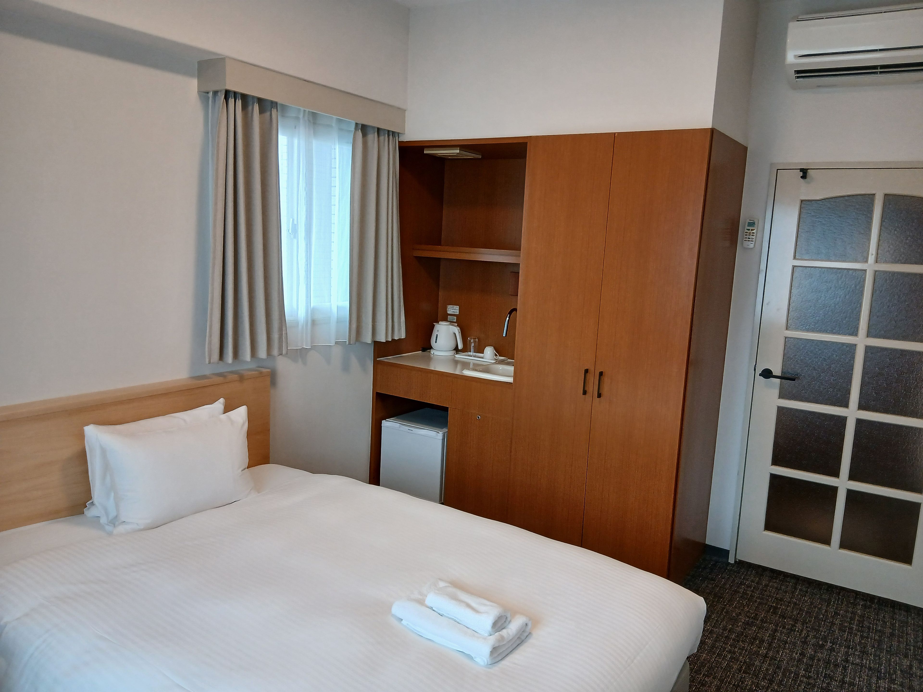 A hotel room with a bed, a wooden wardrobe, a small kitchenette, and a door with frosted glass panels.
