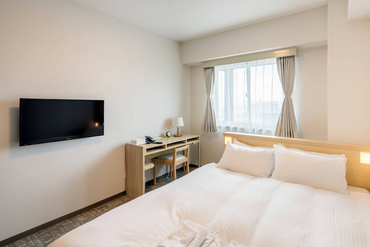 Quarto de hotel com cama, escrivaninha, cadeira, televisão e janela com cortinas.
