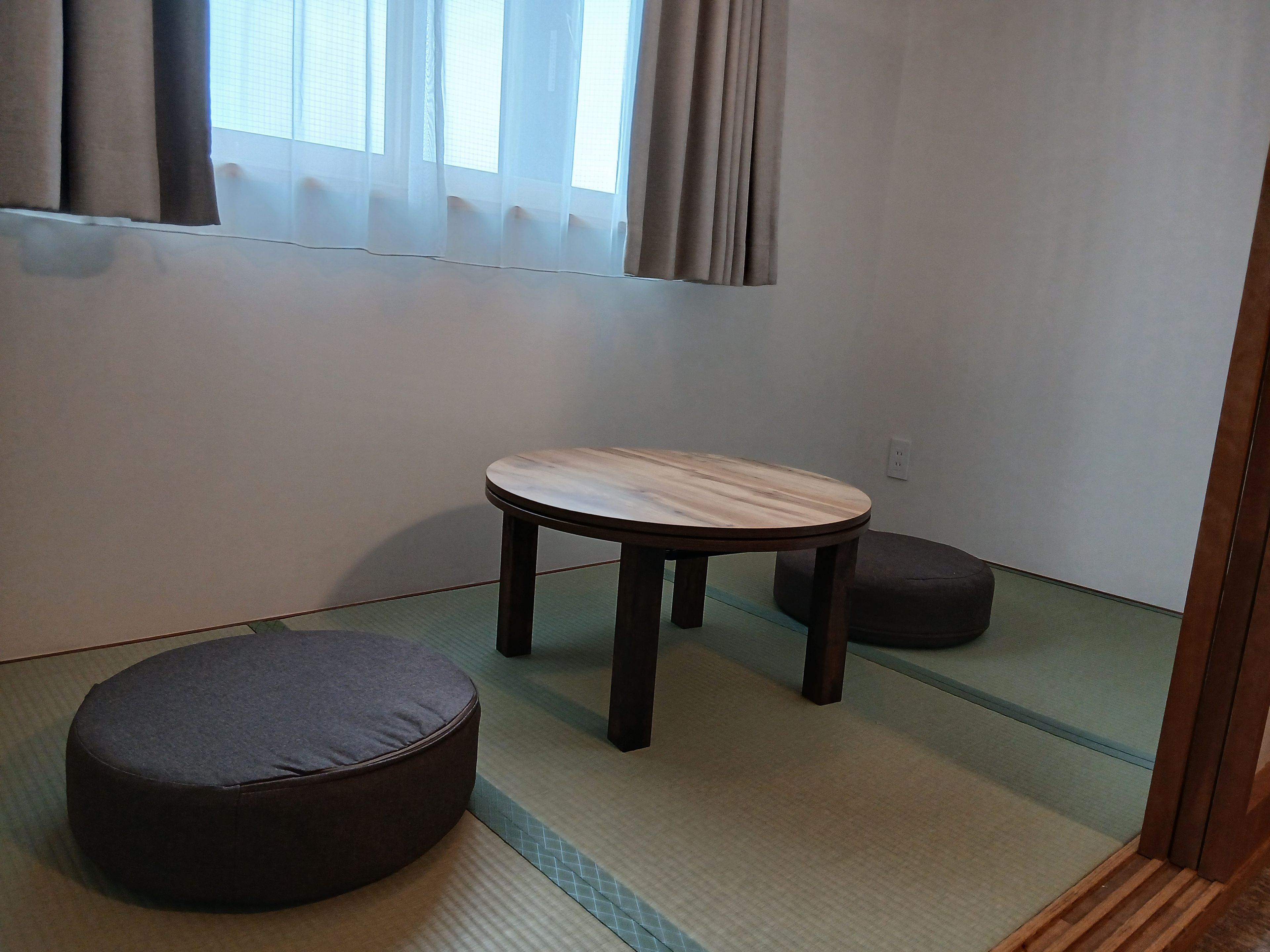Ein Zimmer mit Tatami-Boden, einem niedrigen Holztisch, einem dunklen Kissen und einem Fenster mit Vorhängen.