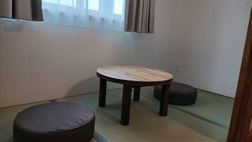 Ein Zimmer mit Tatami-Boden, einem niedrigen Holztisch, einem dunklen Kissen und einem Fenster mit Vorhängen.