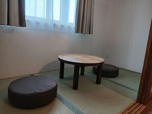 Een kamer met tatami vloerbedekking, een lage houten tafel, een donkere kussen en een raam met gordijnen.