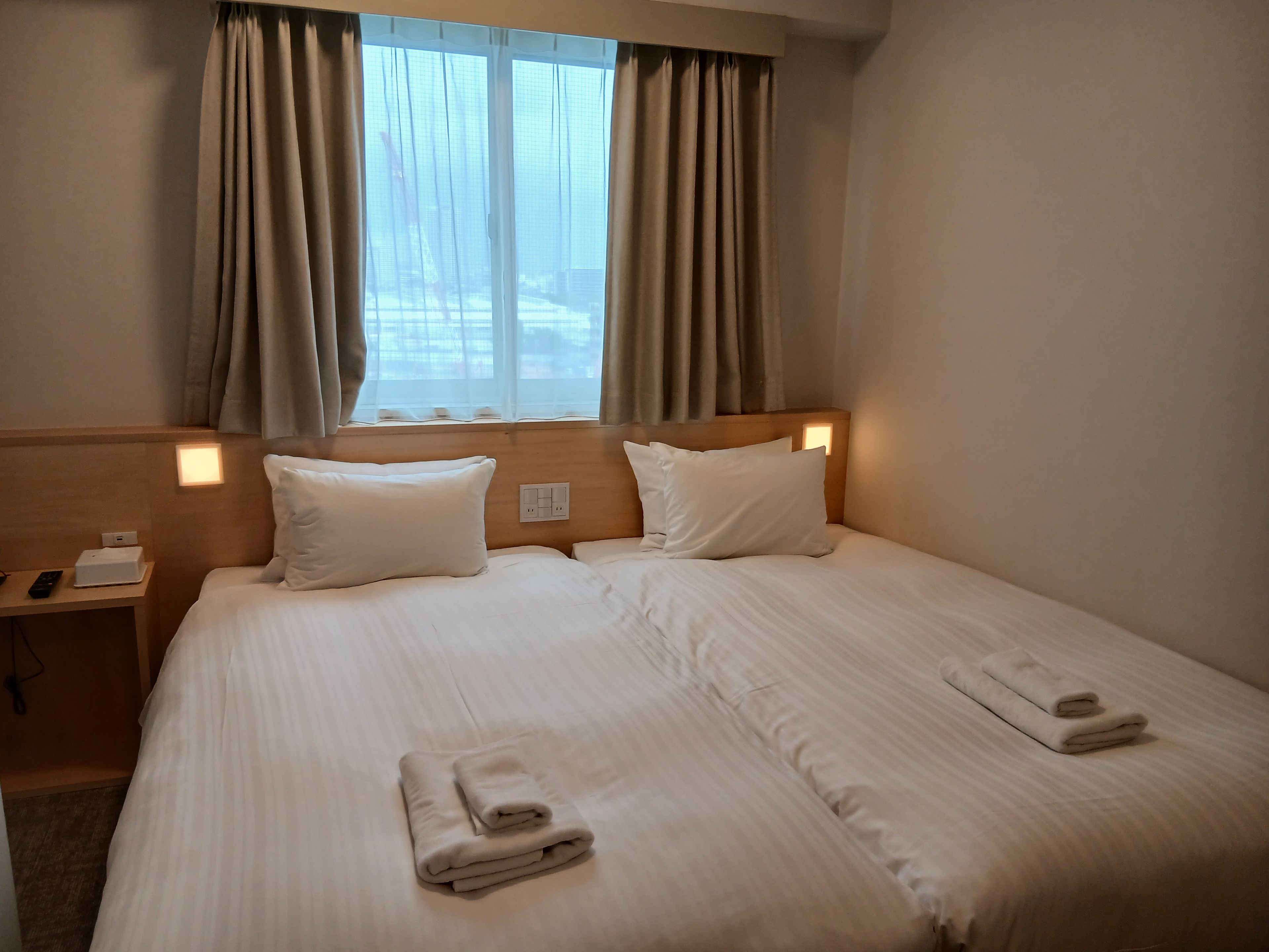 A hotel room with a large bed, two pillows, a nightstand with a phone and a lamp, and a window with curtains.