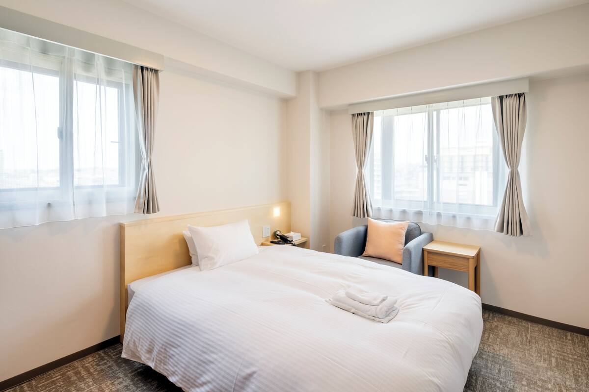 Quarto de hotel com uma cama grande, uma escrivaninha, uma cadeira e duas janelas com cortinas.