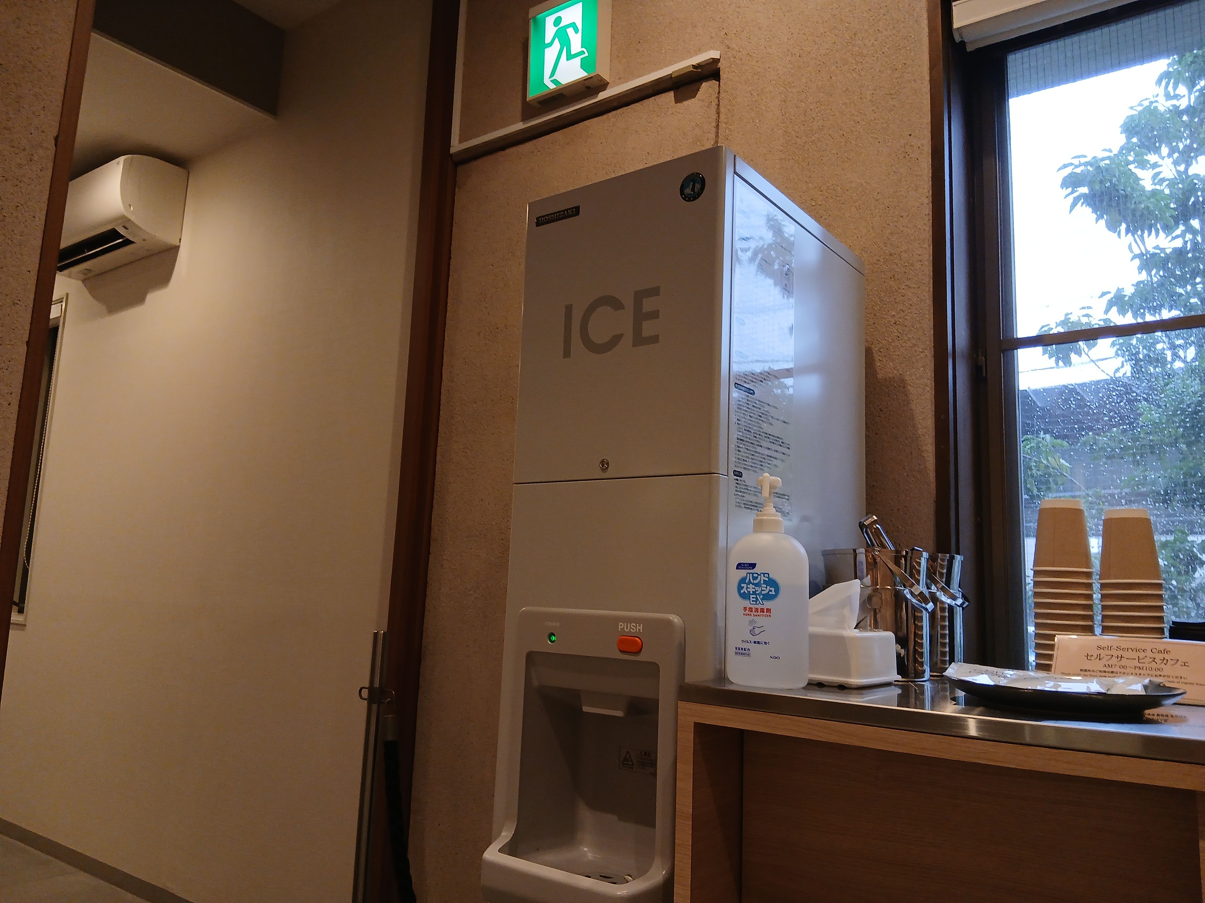 A self-service ice dispenser with a push button, a hand soap dispenser, and a paper towel dispenser in a hotel restroom.