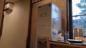 A self-service ice dispenser with a push button, a hand soap dispenser, and a paper towel dispenser in a hotel restroom.