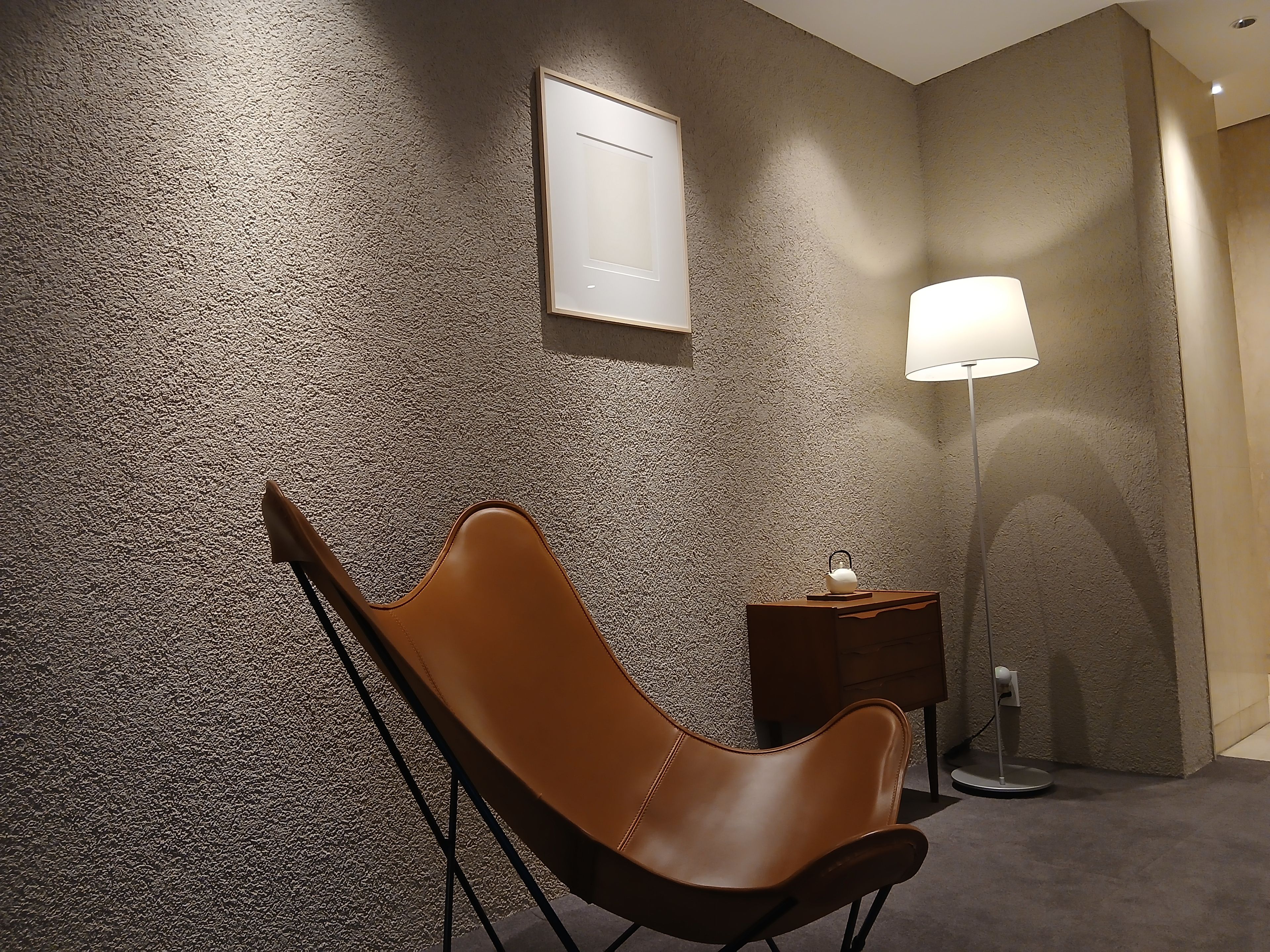 A room with a textured wall, a brown chair, a small wooden side table with a lamp, and a framed picture on the wall.