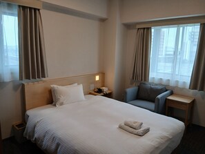 Ein Hotelzimmer mit Bett, Sessel, Nachttisch und einem Fenster mit Vorhängen.