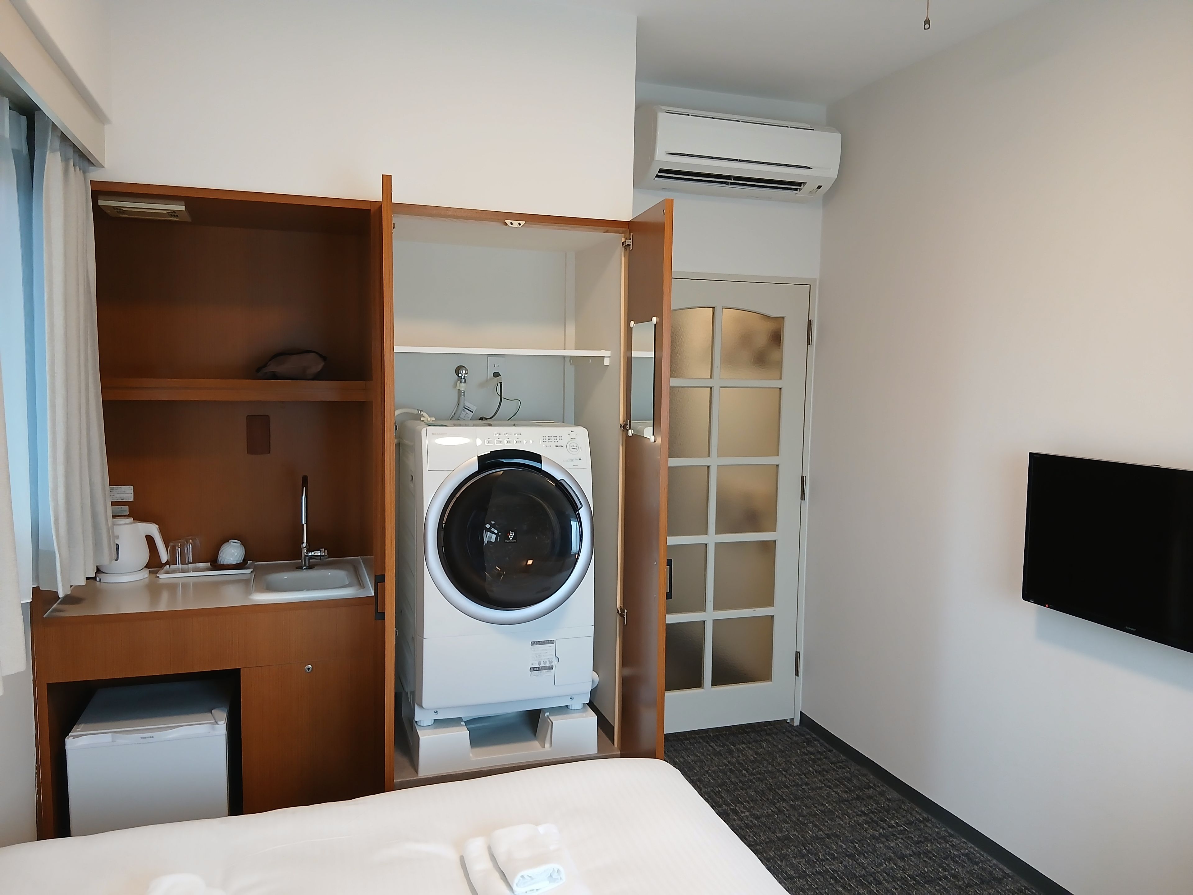 Una habitación de hotel con cama, lavadora, lavamanos pequeño y televisor.