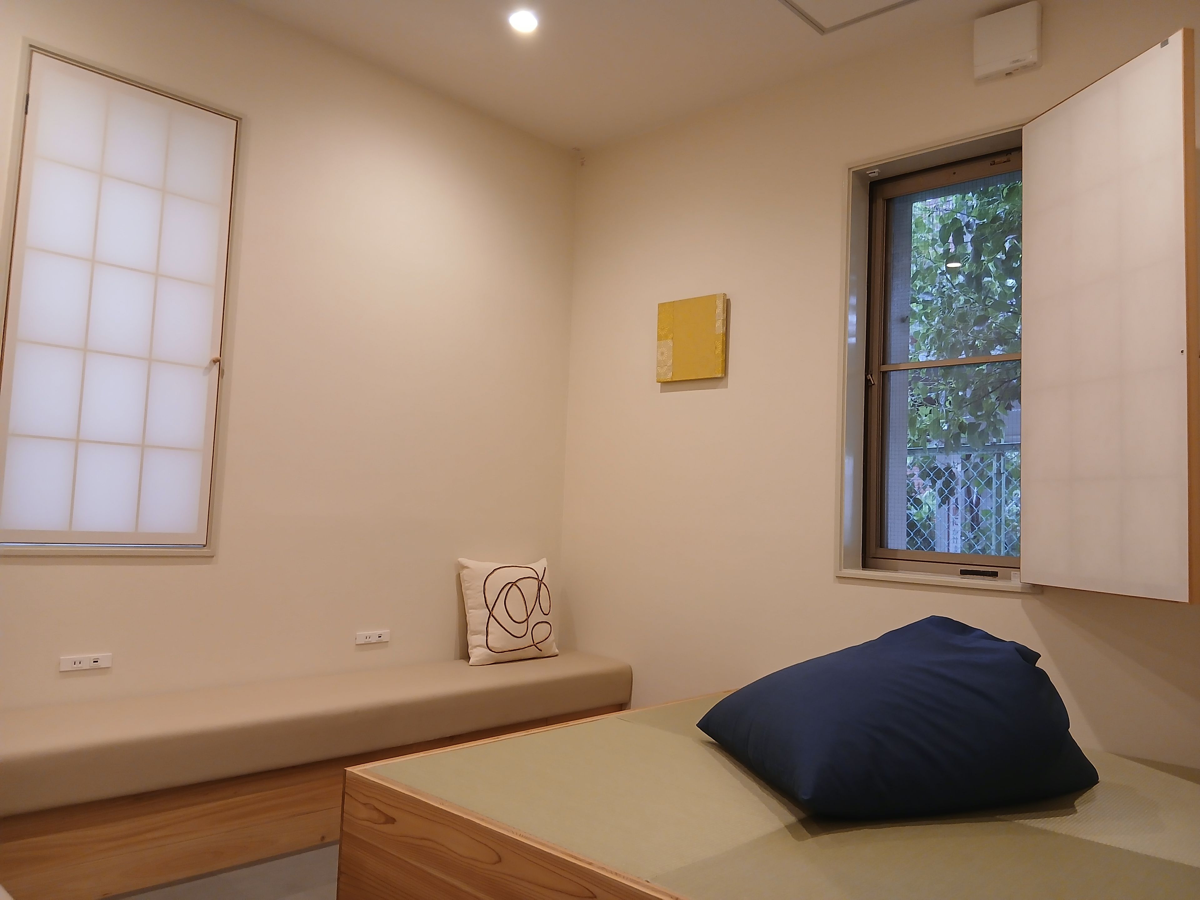 A room with a bed, a window with a view of greenery, a wall-mounted shelf, and a power outlet.