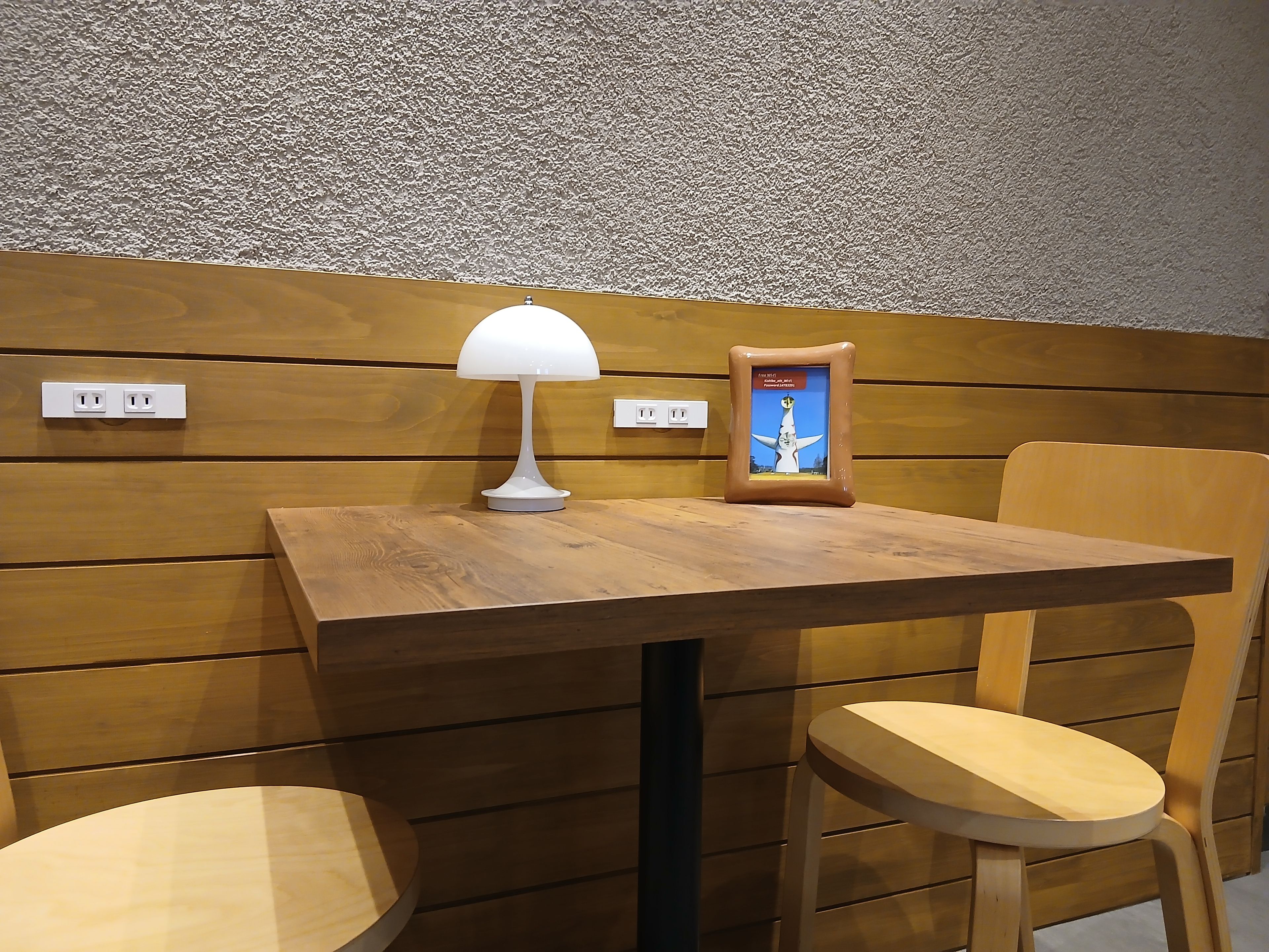 Una mesa de madera con una lámpara blanca y un cuadro enmarcado, dos sillas de madera y una pared con textura y enchufes eléctricos.