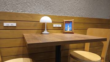 Una mesa de madera con una lámpara blanca y un cuadro enmarcado, dos sillas de madera y una pared con textura y enchufes eléctricos.