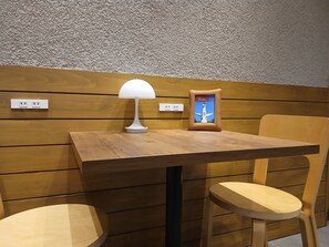 Ein Holztisch mit einer weißen Lampe und einem gerahmten Bild, zwei Holzsessel und eine Wand mit strukturierter Oberfläche und Steckdosen.
