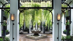 Un patio con una fuente central, rodeado de asientos y plantas en macetas, enmarcado por arcos negros y luces fijadas a la pared.
