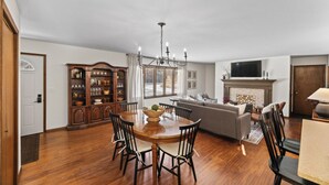 A spacious living room with a fireplace, a dining area with a wooden table and chairs, and a built-in bookshelf. - Remodeled Home on Wooded Nagawicka Lake Channel (Delafield)