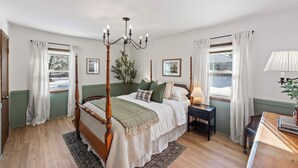 A four-poster bed with a chandelier, a nightstand with a lamp, and a view of the outdoors through the windows. - Remodeled Home on Wooded Nagawicka Lake Channel (Delafield)