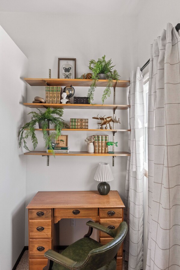 Ein Eckzimmer mit Schreibtisch aus Holz, einem grünen Stuhl und einem Bücherregal mit Dekoration und Pflanzen.