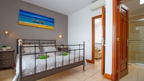 A bedroom with a metal bed frame, a bedside table, a nightstand, a painting of a beach scene, and a door leading to another room. - Casa Yaiza - Three Bedroom Villa, Sleeps 6 (Puerto Calero)