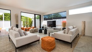A modern living room with two sofas, a coffee table, and a TV. There is a large window providing natural light and a view of greenery outside. - Boutique 5BR Oxygen Pool Villa Samakki 5 (Rawai)