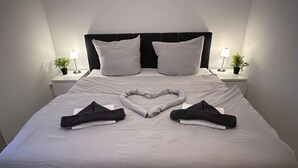 Una cama bien hecha con una toalla dispuesta en forma de corazón, ropa de cama blanca y dos mesitas de noche con lámparas y plantas en macetas.