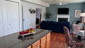 A modern kitchen with wooden cabinets, a granite countertop, and a fruit bowl. There is a dining area with patterned chairs and a table. A fireplace is visible in the background. - Colonial Playland - 5BD, Private Yard, Game Room (Manassas)