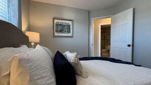 A neatly made bed with white and dark blue pillows, a framed picture on the wall, and a door leading to a bathroom. - Colonial Playland - 5BD, Private Yard, Game Room (Manassas)