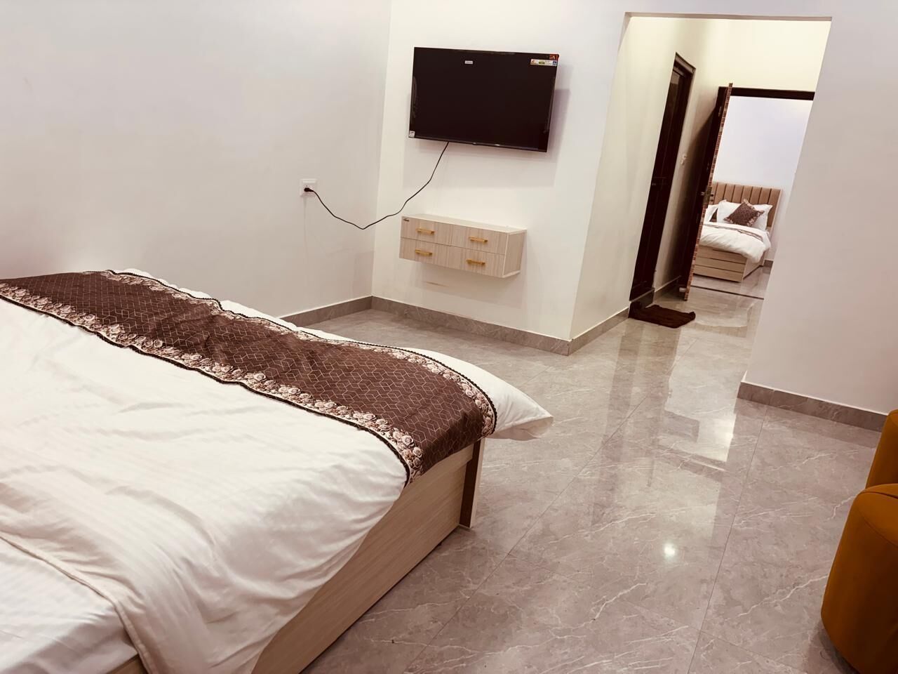 Un dormitorio con cama, televisor montado en la pared, un pequeño mueble de madera y un espejo que refleja una cama en la habitación contigua.