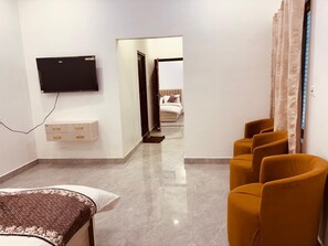 A hotel room with a bed, a TV mounted on the wall, a bedside table, and a seating area with chairs. - Shri Ram Darbar (Faizabad)