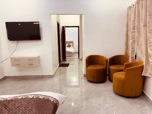 A hotel room with a flat-screen TV mounted on the wall, a bed with a patterned bedspread, two mustard-colored armchairs, and a view into the adjacent room. - Shri Ram Darbar (Faizabad)