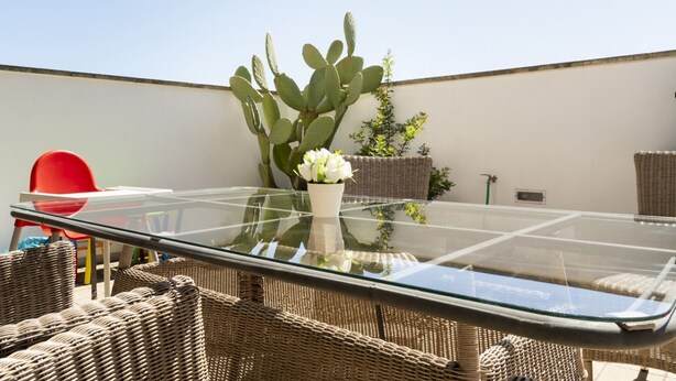 Una mesa con cubierta de vidrio, sillas de mimbre, una silla roja y una planta de cactus.
