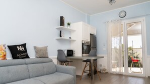 Una sala de estar moderna con un sofá gris, una mesa de comedor con sillas, una zona de cocina con electrodomésticos y una puerta corrediza de vidrio que da a un área exterior.