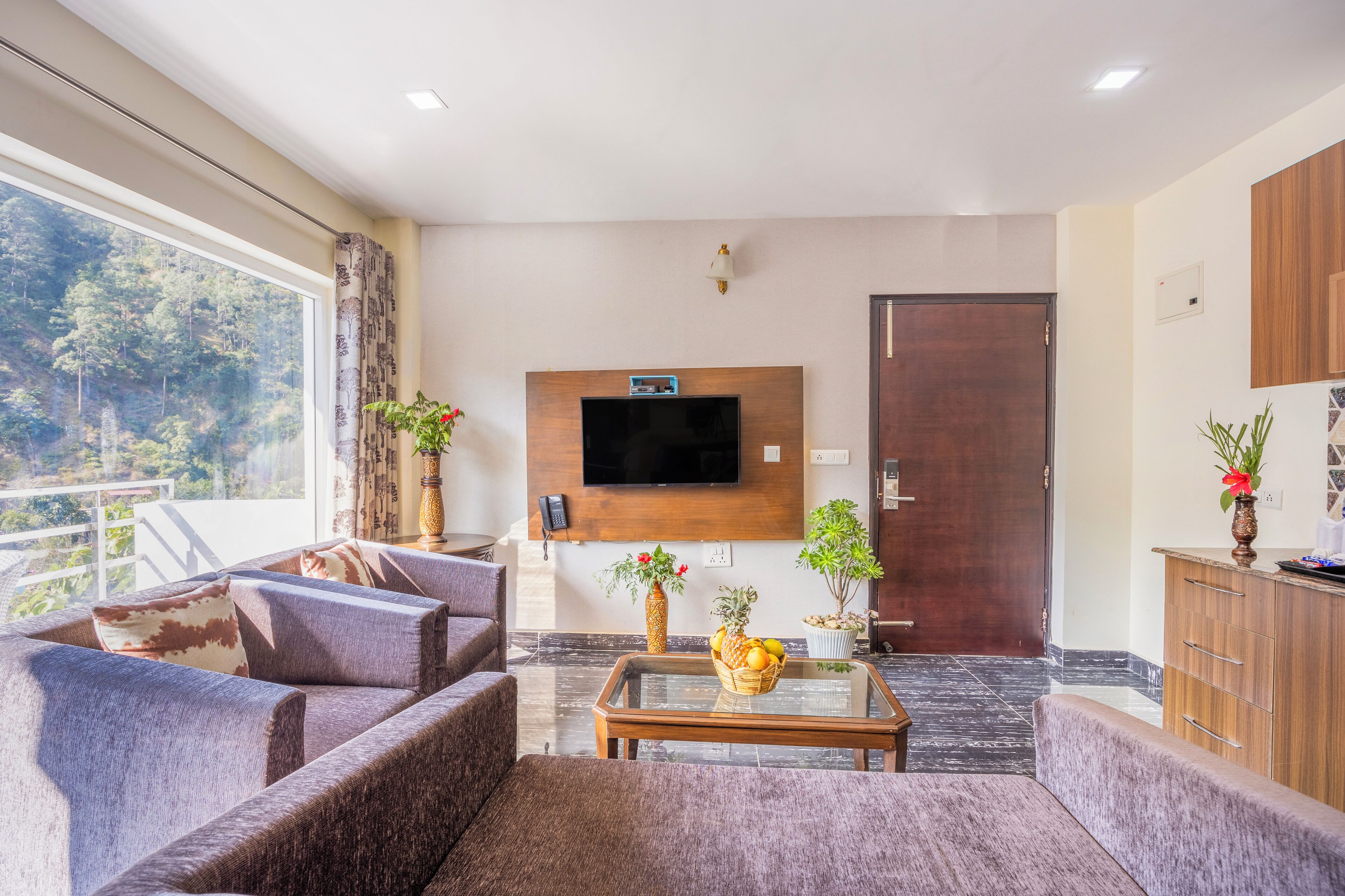 Un salon moderne comprenant un ensemble de canapés, un téléviseur à écran plat fixé au mur, un meuble en bois et une grande fenêtre donnant sur une zone boisée.