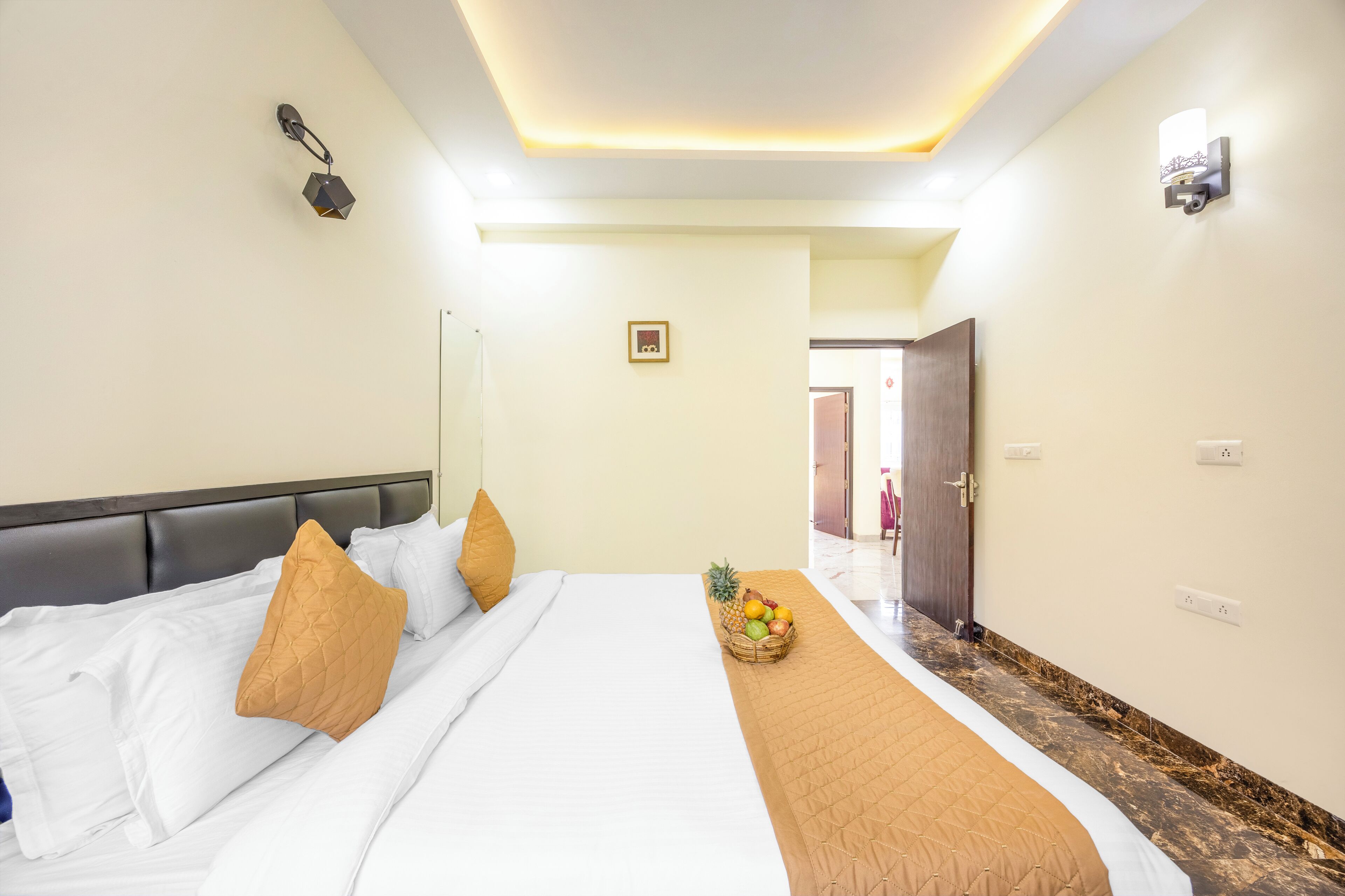 A hotel room with a large bed, a fruit basket on the bed, a brown throw blanket, and a modern ceiling light fixture.