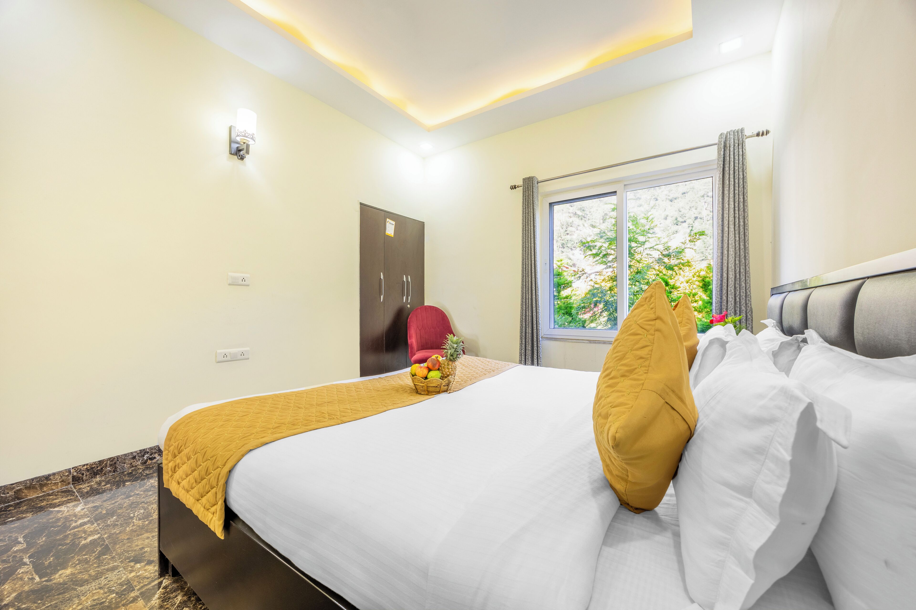 A hotel room with a bed, a red chair, a fruit basket, and a window with a view of greenery.