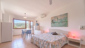 A bedroom with a bed, a desk, and a chair. There is a window with a view of the outdoors.