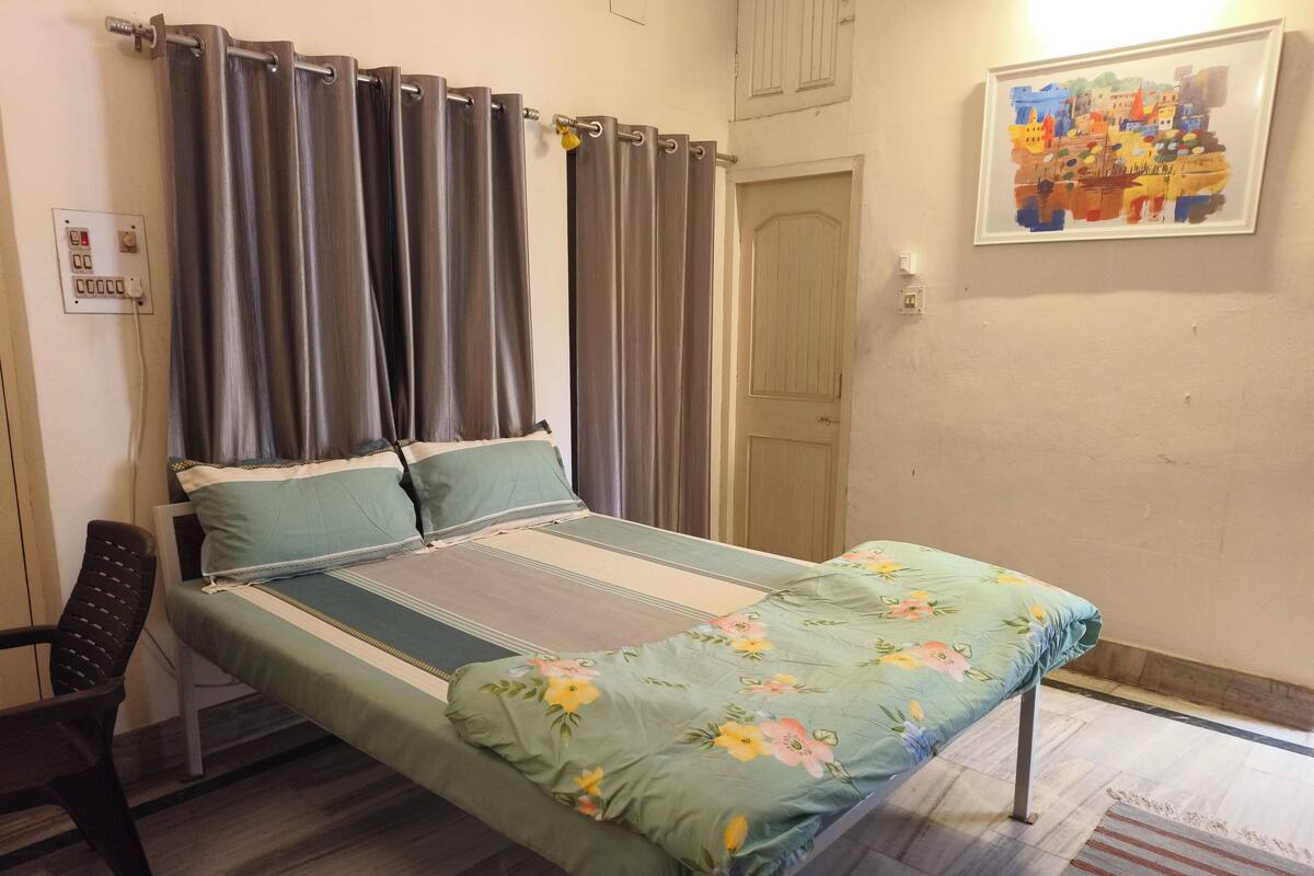 A single bed with a floral bedspread, a chair, a wooden door, and a painting on the wall.