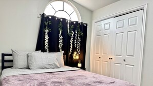 A bedroom with a bed, a window with curtains, a nightstand, and a white door.
