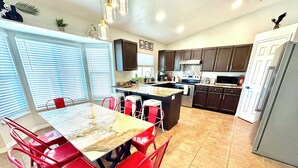 A kitchen with a dining area, featuring a central island, red chairs, and a large window with blinds.
