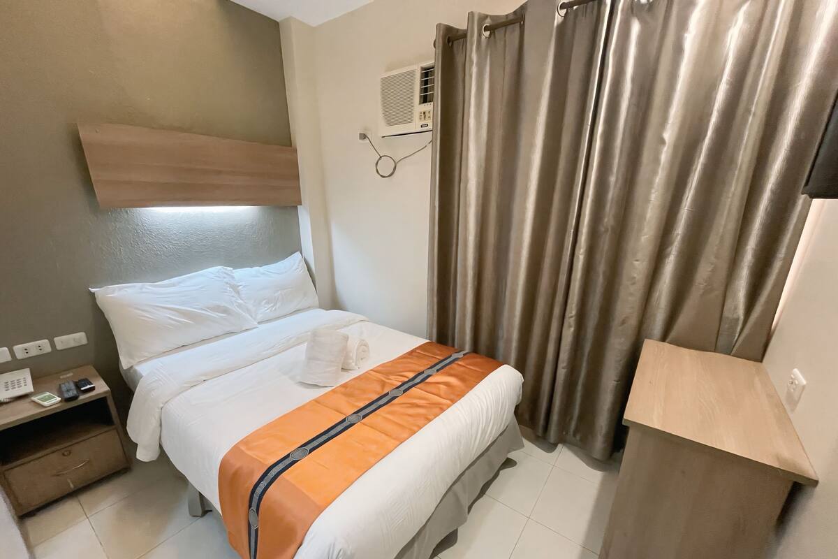 Habitación de hotel con cama, mesita de noche con teléfono y controles remotos, aire acondicionado en la pared y ventana con cortinas.