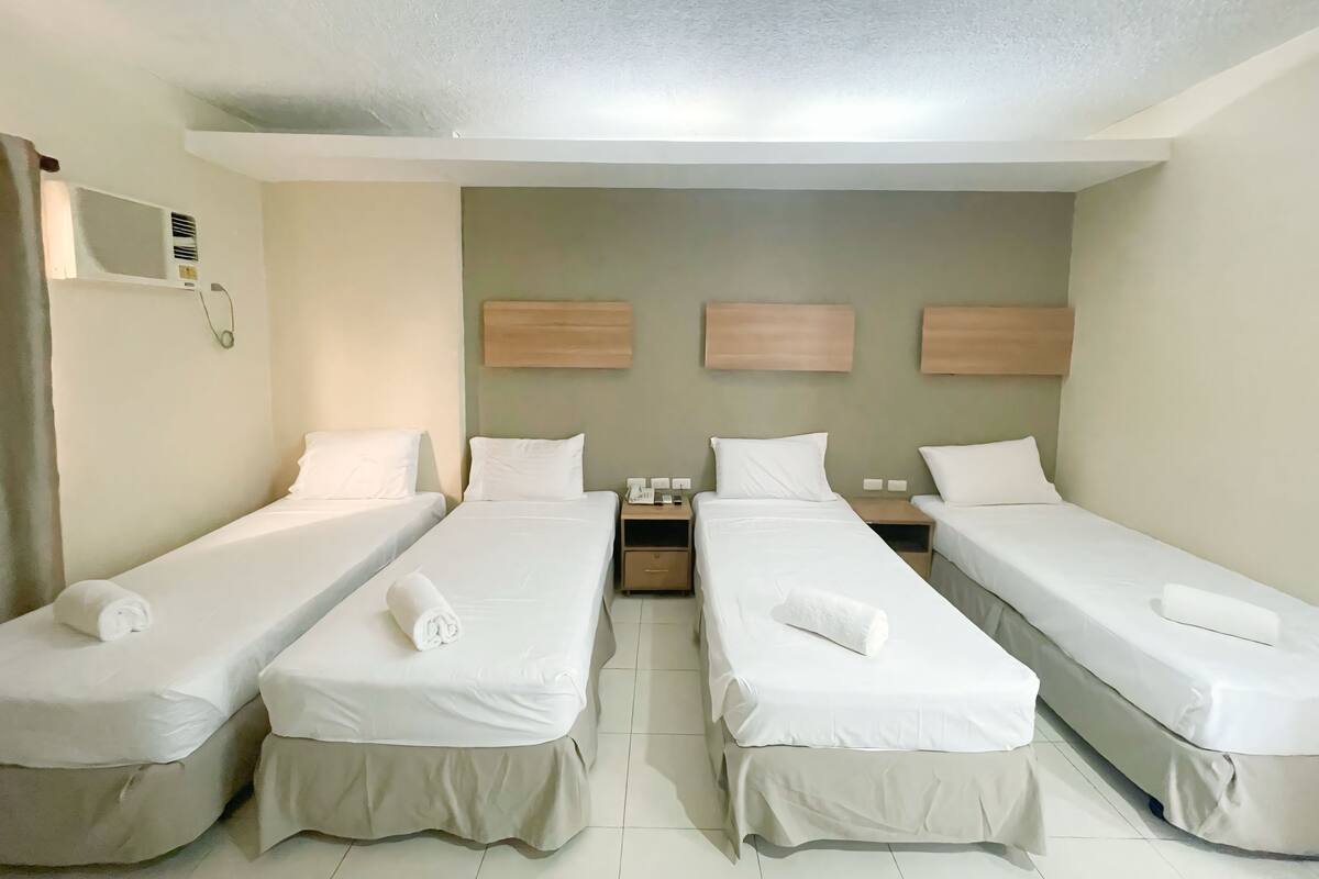 Habitación de hotel con cuatro camas individuales, cada una con una colcha blanca y una toalla enrollada. Hay dos mesitas de noche con lámparas y una pequeña mesa entre las camas. La habitación presenta una decoración en tonos neutros, con paredes color beige y techo blanco.