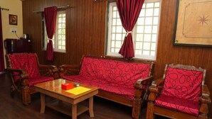 Un salón comedor con muebles de madera, un sofá, dos sillones, una mesa de centro de madera y una mesita con un objeto rojo y un papel amarillo.