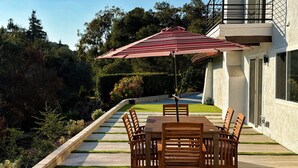 Eine Terrasse mit Tisch, Stühlen und einem Sonnenschirm, mit Blick auf einen Garten und ein Haus.