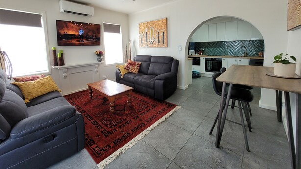 A modern living room with a grey sofa, a red rug, and a wooden coffee table. A kitchen with white cabinets and a herringbone backsplash is visible through an archway.