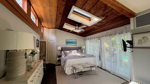 A bedroom with a large bed, wooden ceiling, skylight, bedside table, lamp, and a view of the outdoors.